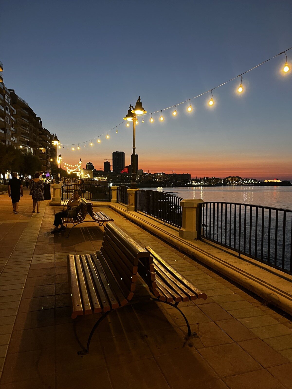Sliema seafront promenade at sunset, with string lights and benches along the water